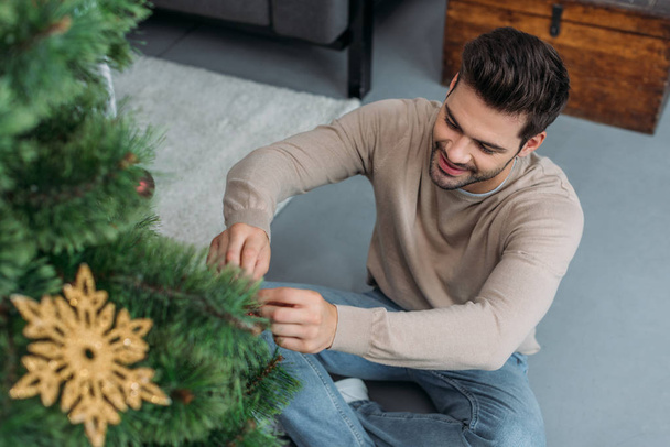 Hochwinkel-Ansicht des lächelnden gutaussehenden Mannes, der Weihnachtsbaum mit Kugeln dekoriert und zu Hause auf dem Boden sitzt - Foto, Bild