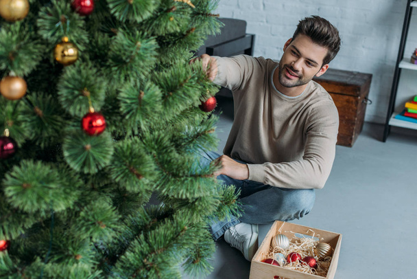 красивый мужчина украшает рождественскую елку безделушками и сидит дома на полу
 - Фото, изображение