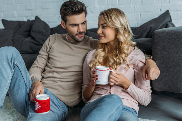 petit ami souriant et petite amie tenant des tasses de cappuccino et assis sur le tapis dans le salon
 - Photo, image