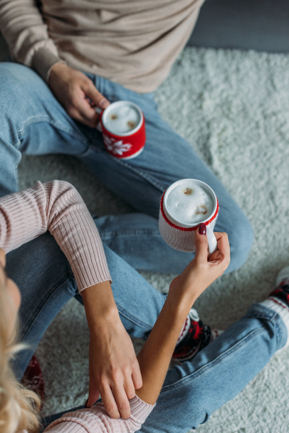 cropped image of couple sitting on carpet and holding cups of cappuccino at home, christmas concept - Photo, image
