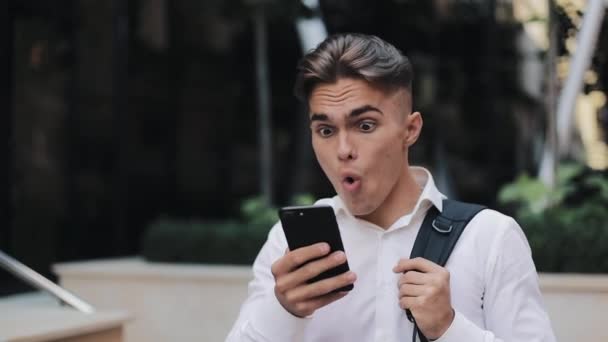 Young surprised businessman looking shocked at smartphone screen checking notification bad news. Man with backpack walking near modern office building. - Footage, Video