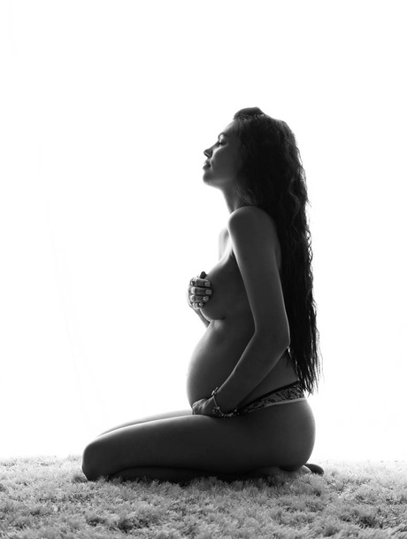 Intimate portrait of beautiful pregnant woman covers herself with hands while posing topless at home - black and white image - Foto, Imagem