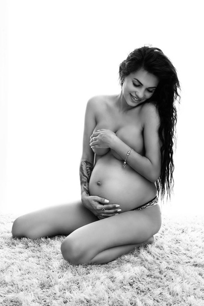 Intimate portrait of beautiful pregnant woman covers herself with hands while posing topless at home - black and white image - Foto, Bild