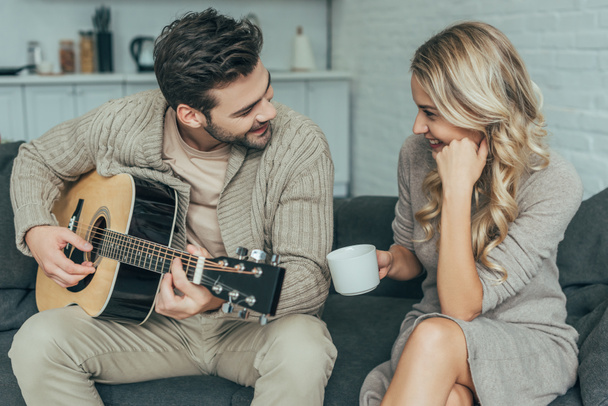happy young man playing guitar for girlfriend at home while she sitting on couch with coffee - Φωτογραφία, εικόνα