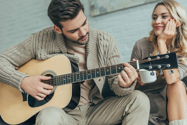 handsome young man playing guitar for girlfriend at home while she sitting on couch with coffee - Foto, Imagem