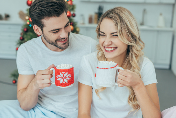 смеющаяся молодая пара с чашками какао, сидящая в постели дома на Рождество
 - Фото, изображение