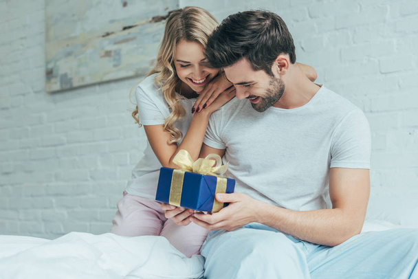 beautiful young woman presenting gift to boyfriend in bed at home - Foto, immagini