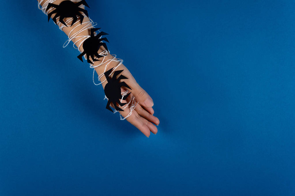 Feminine hand wrapped in white web with black paper spiders on blue background. Halloween concept. Paper cut  style. Top view - Фото, изображение