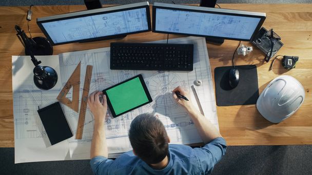 Top View of Architectural Engineer Draws on His Blueprints, Compares with Tablet Computer with Green Screen, Using Desktop Computer Also. His Desk is Full of Useful Objects and Evening Sun. - Foto, imagen