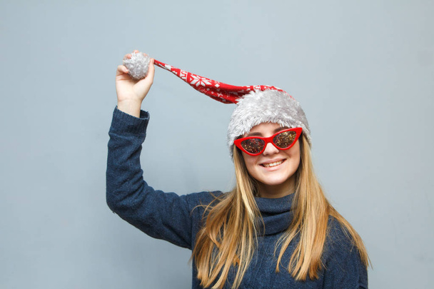 Christmas girl in Santa hat and sunglasses stylized sixties  smiles on grey background - Photo, Image