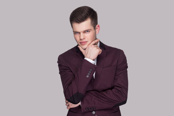 Portrait of handsome serious young man in violet suit and white shirt touching his chin and looking at camera with thoughtful face on grey background. - Valokuva, kuva