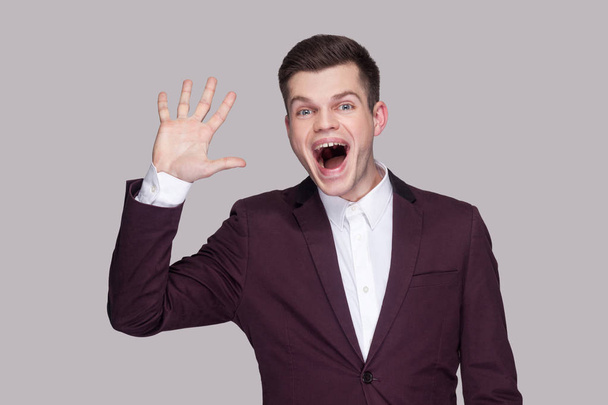 Portrait of funny handsome young man in suit and white shirt looking at camera with amazed face and greeting gesture on grey background. - Fotografie, Obrázek