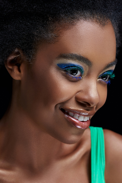 portrait of smiling african american model with bright blue eyes shadows isolated on black - Fotografie, Obrázek