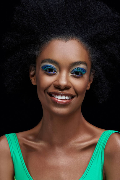 portrait of smiling african american model with bright blue eyes shadows isolated on black - 写真・画像