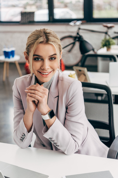 beautiful young businesswoman sitting at workplace and smiling at camera - Foto, Imagen