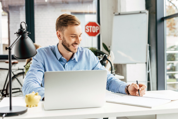 sorridente jovem empresário usando laptop e tomar notas no local de trabalho
   - Foto, Imagem