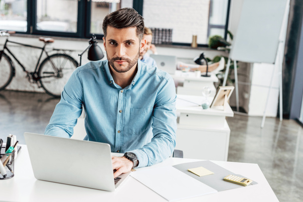 handsome young businessman using laptop and looking at camera in office  - Fotografie, Obrázek