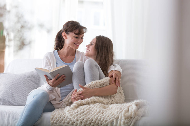 Happy mother reading book to smiling daughter while relaxing at home - Foto, Bild
