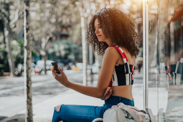 Charming African-American female is sitting on the bench inside of urban bus stop and having a video call via her smartphone; a dazzling woman with curly Afro hair with cellphone is waiting for a bus - Photo, image