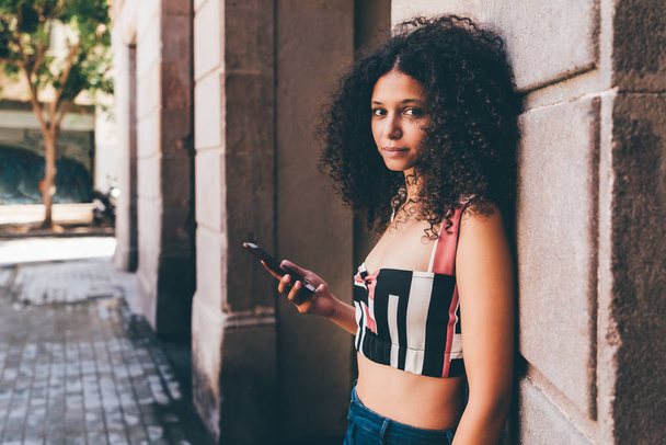 Portrait of a young beautiful Moroccan female tourist leaning against the wall outdoors and holding her smartphone; charming curly African-American woman with the cell phone on the street of Barcelona - Foto, Imagem