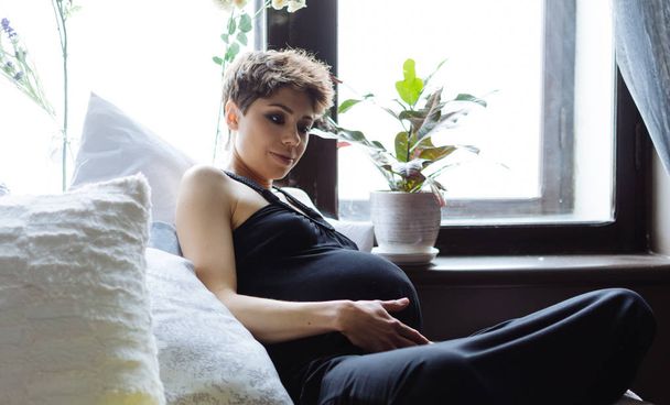 Future mother sits on a bed. The pregnant woman , in a black dress - Valokuva, kuva