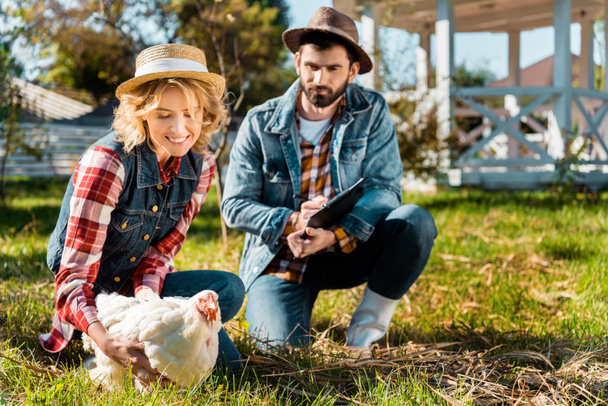 selective focus of woman holding chicken while her boyfriend writing in clipboard at farm - Фото, изображение