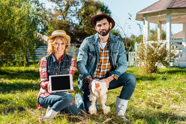 mujer mostrando tableta digital con pantalla en blanco mientras su novio sostiene pollo en la granja
 - Foto, imagen