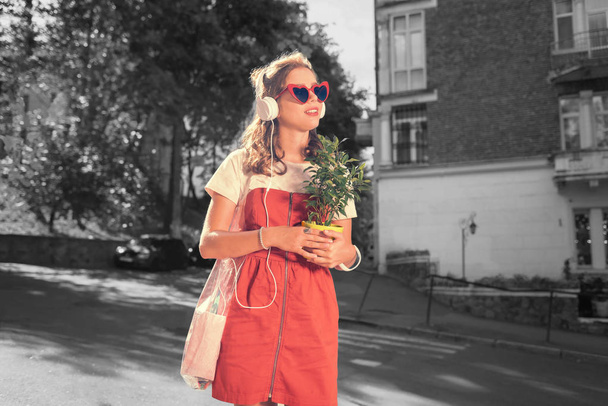Woman with plant. Beautiful dark-haired woman listening to music in earphones holding home plant on her way home - Foto, afbeelding
