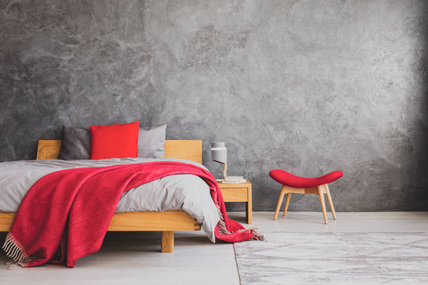 Red pillow and blanket on comfortable bed with grey bedding in simple bedroom with copy space on the empty concrete wall, real photo - Fotografie, Obrázek