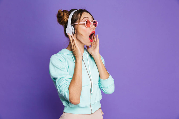 Portrait of an excited young girl with bright makeup over violet background, listening to music with headphones - Foto, afbeelding