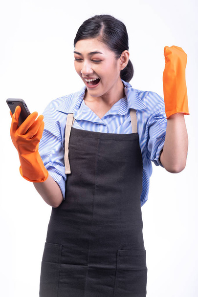 House cleaning service woman isolated in white. Asian young woman with gloves, Using mobile phone, big win pose. Online house cleaning service concept. - Фото, изображение