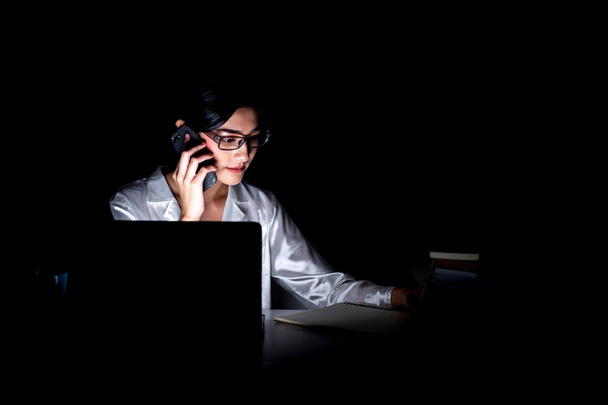 Woman working late at her home office. Beautiful asian woman talking on phone and looking at her computer, talking to customer, taking notes, wearing glasses. Modern global business concept. - Foto, Imagen