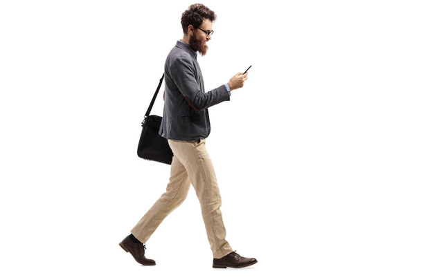 Full length profile shot of a man walking and looking into a mobile phone isolated on white background - Zdjęcie, obraz