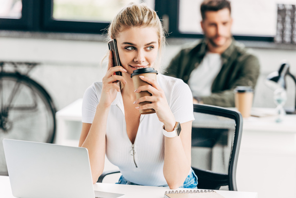 beautiful young woman talking by phone and drinking coffee at office with colleague on background - Photo, Image