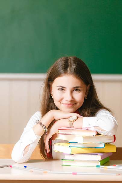 schoolgirl in school uniform sitting at her desk with books and pencils on the background of a green board. - Foto, afbeelding