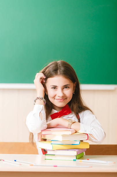 colegiala en uniforme escolar sentada en su escritorio con libros y lápices en el fondo de un tablero verde
. - Foto, Imagen