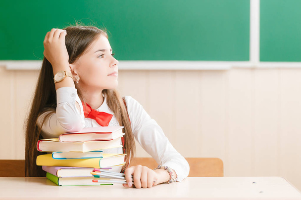 Schulmädchen in Schuluniform sitzt an ihrem Schreibtisch mit Büchern und Bleistiften auf dem Hintergrund einer grünen Tafel. - Foto, Bild