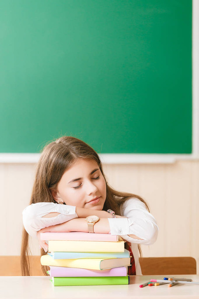 schoolgirl in school uniform sitting at her desk with books and pencils against the background of a green blackboard and sleeping - Foto, Imagen