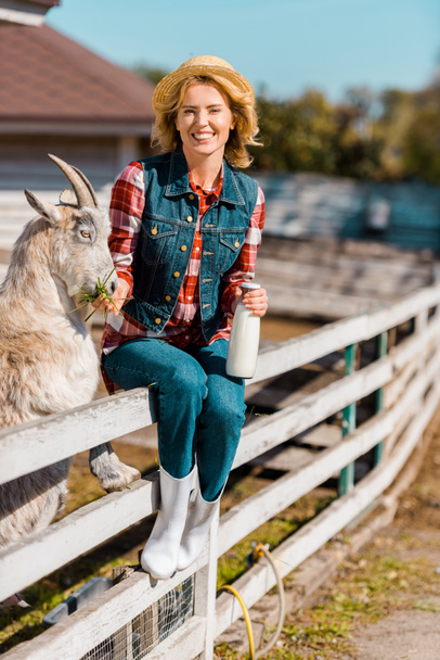 усміхнена жінка-фермерка з пляшкою молока, що сидить на дерев'яному паркані і годує козла на ранчо
 - Фото, зображення