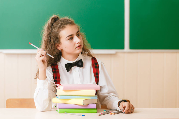школьница в школьной форме сидит за своим столом с книгами и карандашами на фоне зеленой доски
. - Фото, изображение