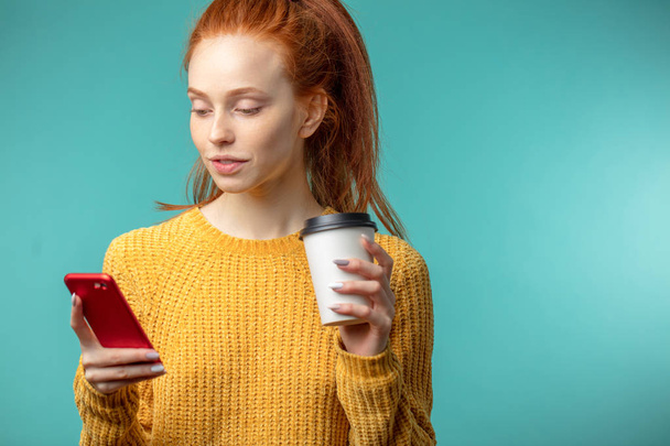 Jonge roodharige vrouw met koffie en smartphone geïsoleerd op blauwe achtergrond - Foto, afbeelding