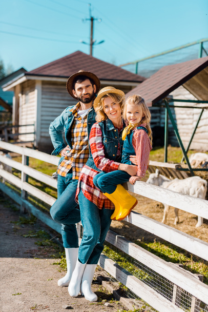 familia campesina sonriente con una hija pequeña sentada en una cerca de madera en la granja
  - Foto, Imagen