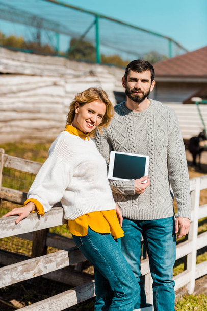 sonriente pareja de agricultores mostrando tableta digital con pantalla en blanco cerca de valla de madera en la granja
 - Foto, Imagen