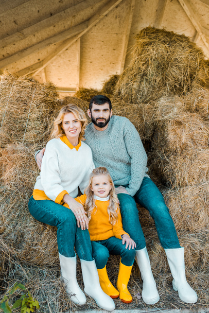 cheerful farmer family with daughter sitting on hay stacks at ranch - Photo, Image