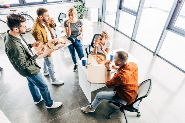 korkean kulman näkymä ryhmä nuoria yrittäjiä, jotka syövät pizzaa lounaaksi yhdessä työskennellessään käynnistyksen toimistossa
 - Valokuva, kuva