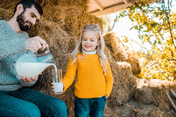 adult female farmer pouring milk to little daughter near hay stacks at ranch - Photo, Image