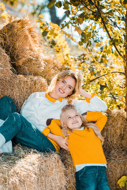 smiling adult woman posing with little daughter on hay stacks at farm - Photo, Image