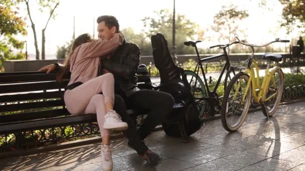 A beautiful caucasian couple sit embracing on a park bench. Portrait of a couple in love on a bench in the park and having fun together. Black guitar case and bike next to them - Video