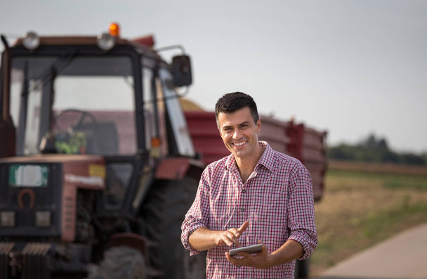 Granjero guapo con la tableta de pie delante del tractor con remolques en el campo
 - Foto, Imagen