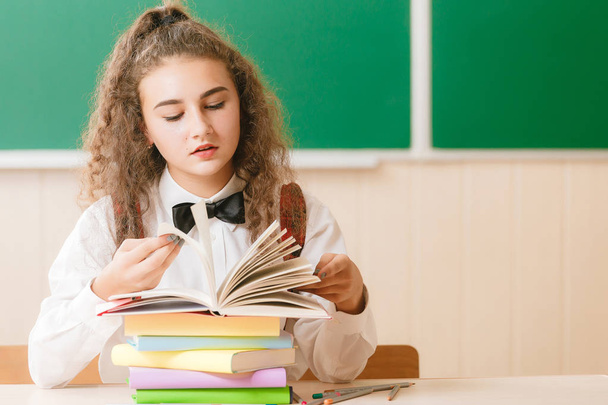 colegiala en uniforme escolar sentada en su escritorio con libros y lápices en el fondo de un tablero verde
 - Foto, Imagen